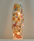 A tall, narrow white decorative lamp or vase with a warm internal light, decorated with colorful painted flowers and butterflies, set against a soft white  background.