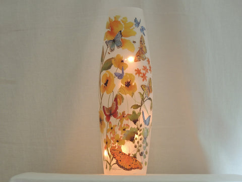 A tall, narrow white decorative lamp or vase with a warm internal light, decorated with colorful painted flowers and butterflies, set against a soft white  background.