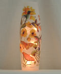 A tall, narrow frosted glass candle vase lamp featuring hand-painted hummingbirds, butterflies, and yellow flowers, illuminated from within against a neutral background.