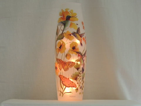 A tall, narrow frosted glass candle vase lamp featuring hand-painted hummingbirds, butterflies, and yellow flowers, illuminated from within against a neutral background.