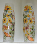 Slender frosted glass vase unlit, decorated with colorful illustrations of yellow flowers, hummingbirds, butterflies, and green leaves, set against a plain white background.