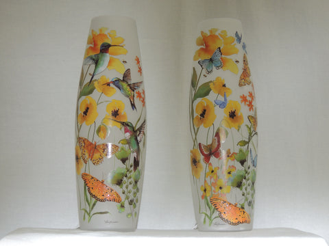 Slender frosted glass vase unlit, decorated with colorful illustrations of yellow flowers, hummingbirds, butterflies, and green leaves, set against a plain white background.