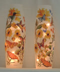 Tall frosted glass lamp vase decorated with colorful painted flowers, hummingbirds, and butterflies, glowing with warm internal lights against a neutral draped backdrop.